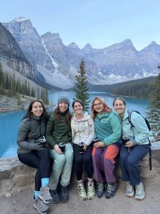 5 women posing on a wall in front of turquoise Moraine Lake, Banff National Park.